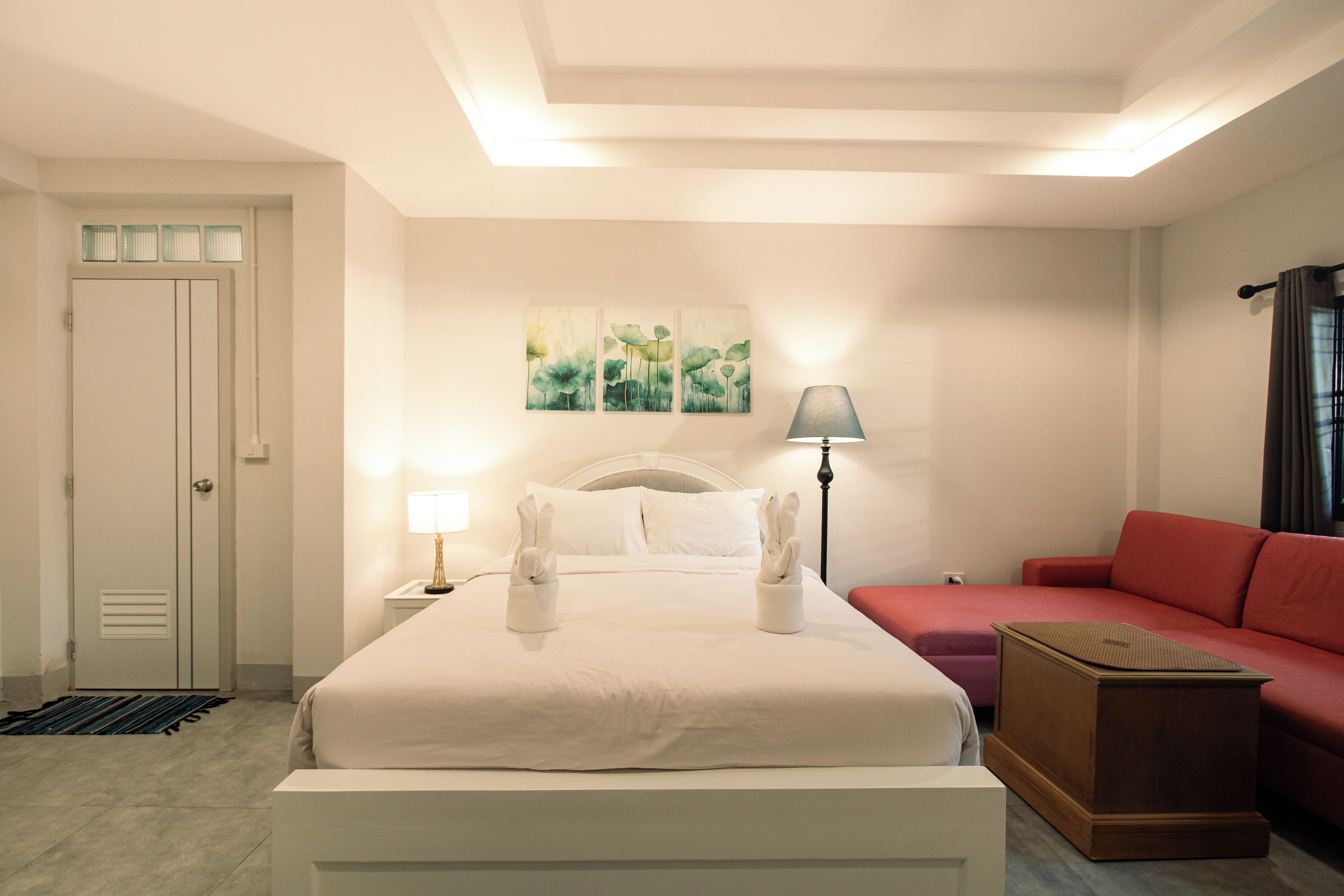 A hotel room with a bed, a red sofa, a nightstand with a lamp, a painting on the wall, and a door with a window.
