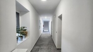 A narrow, well-lit hallway with white walls and grey tiled flooring. There are potted plants on the left side and a door at the end. - Rooms Burnum (Biskupija)