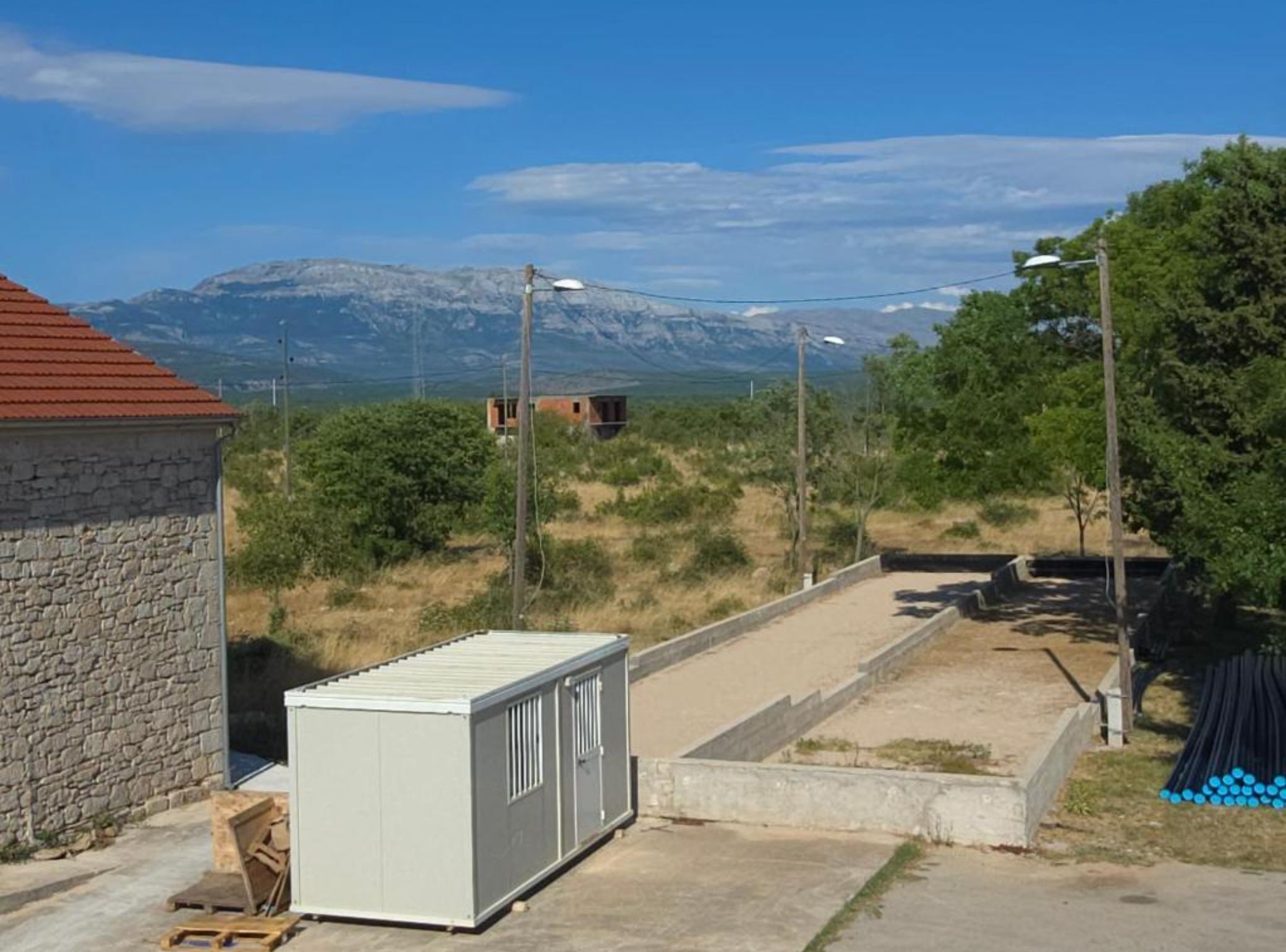 A rural setting with a stone building, a small shed, and a paved area with a metal fence.