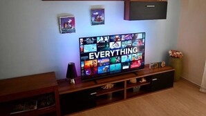 A living room with a flat-screen TV on a wooden stand, surrounded by shelves with decorative items and a few framed pictures. - Apartment Zara (Marina)