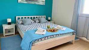 A bedroom with a teal wall, a bed with patterned bedding, a small bedside table with a lamp, and a window with curtains. - Apartment Zara (Marina)