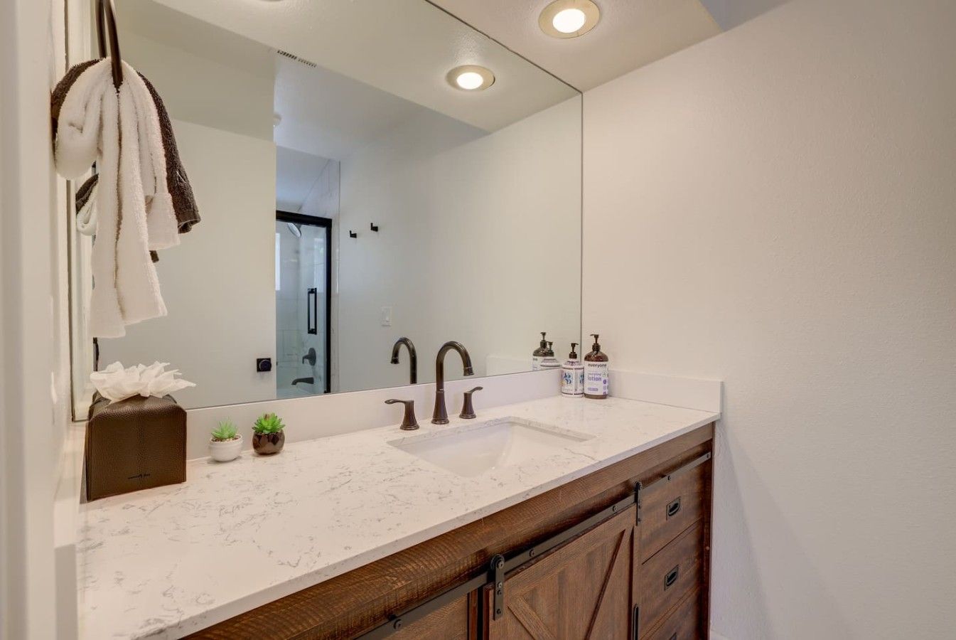 A bathroom with a large mirror, a white sink, two faucets, a towel rack, a tissue box, and a small plant.
