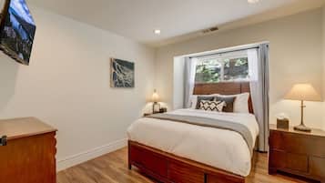 A bedroom with a wooden bed, bedside lamps, a flat-screen TV mounted on the wall, and a window with curtains.
