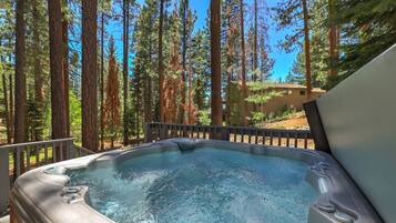 A hot tub surrounded by tall pine trees, with a wooden deck and a building in the background.