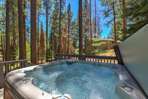 A hot tub surrounded by tall pine trees, with a wooden deck and a building in the background. - Family Retreat | Master Suites-Hot Tub | Sleeps10 (Stateline)