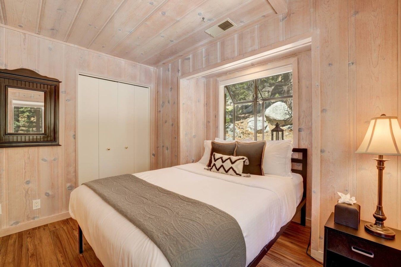 A bedroom with a bed, a window with a view of rocks and trees, a bedside table with a lamp, and a wardrobe.