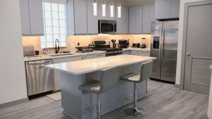 A modern kitchen with grey cabinets, a stainless steel refrigerator, a microwave, and a large island with bar stools. - Luxury Home - near NRG, Medical Center, Downtown & Galleria (5 - 15 minutes) (Houston)
