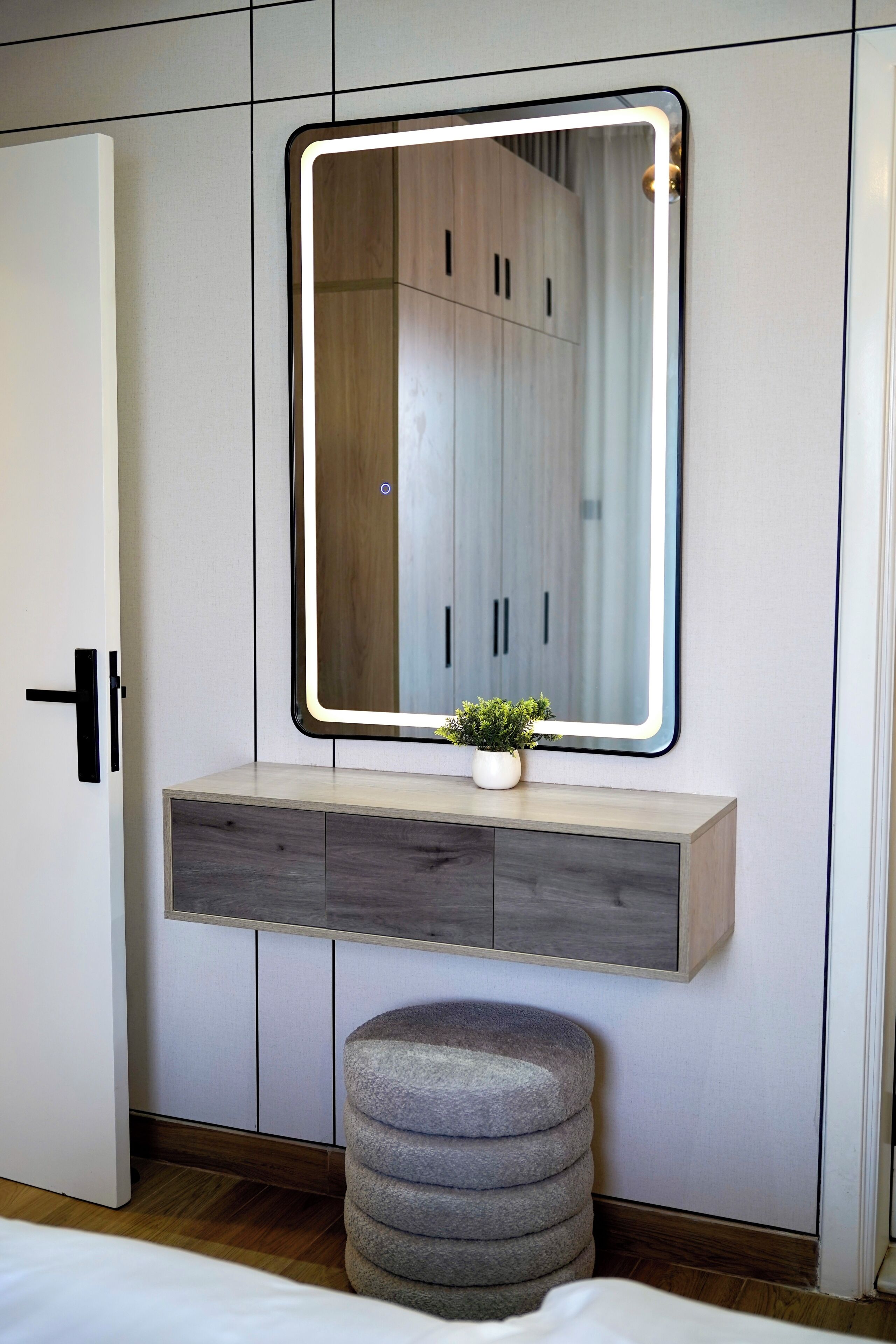 Une chambre avec un grand miroir, une table de chevet agrémentée d’une plante et un pouf gris.
