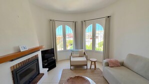 Una sala de estar con chimenea, un sofá, una butaca y una mesa de centro. Hay dos ventanas con cortinas y vistas a edificios.