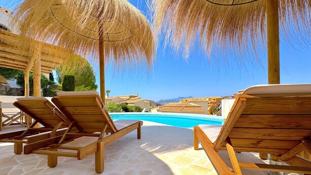 Zona de piscina con sillones de madera y sombrillas de paja, piscina azul cristalina y vista a edificios y montañas al fondo.