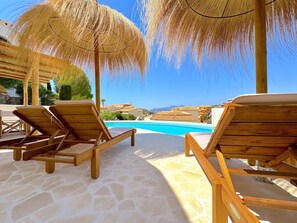 A poolside area with wooden lounge chairs and thatched umbrellas, a clear blue pool, and a view of buildings and mountains in the background. - Villa Maeva - Benitachell, Costa Blanca (Benitachell)