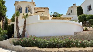 Un edificio blanco con fachada curva, una pequeña torre con ventana y un camino de piedra bordeado de flores rosadas.