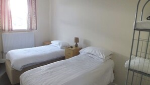 A room with two single beds, a bedside table with a lamp, a radiator, and a window with curtains. - OYO Lodge Bournemouth Standard Family of 3 Room (Bournemouth)