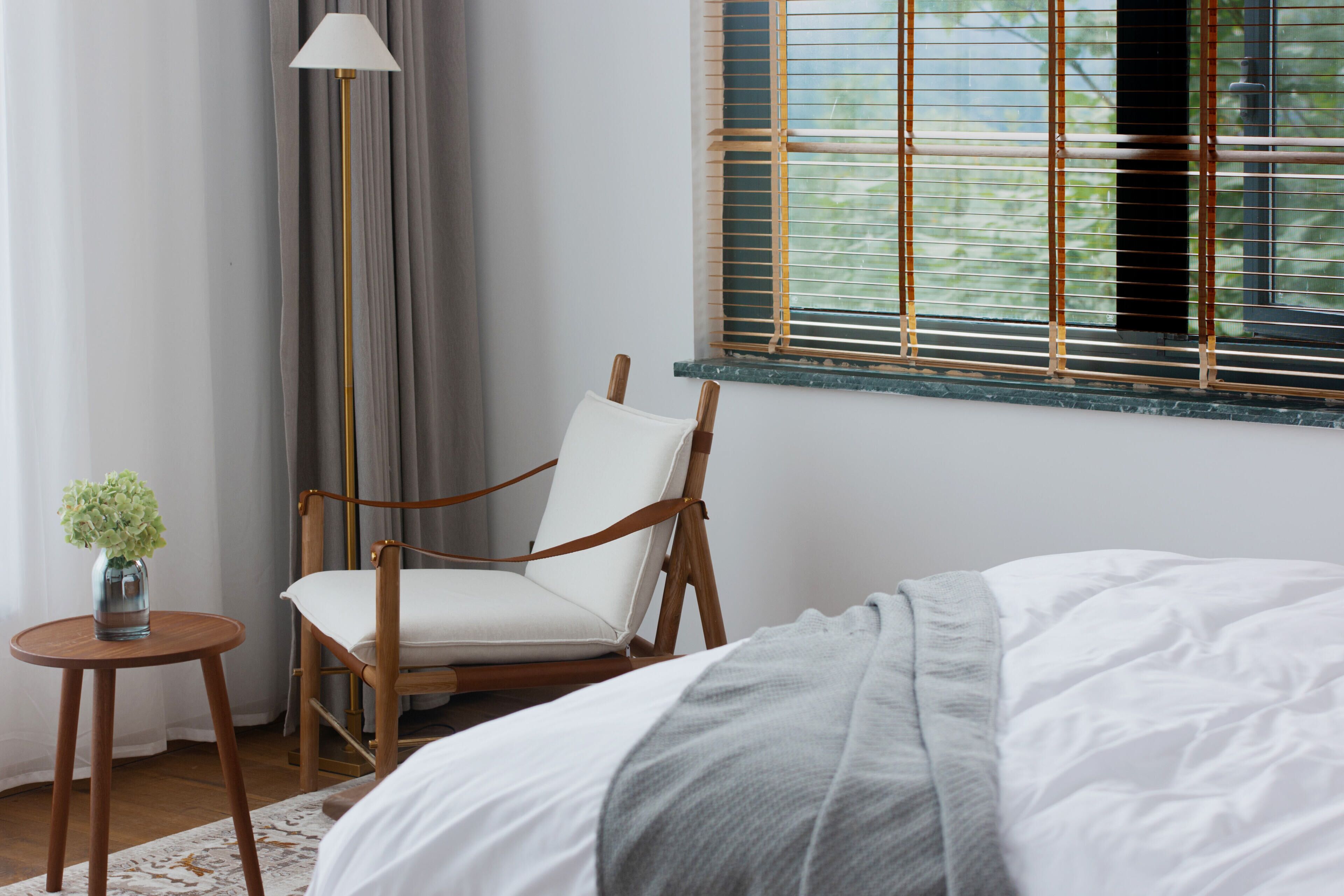 A bedroom with a bed, a chair, a small table, a lamp, and a window with blinds.