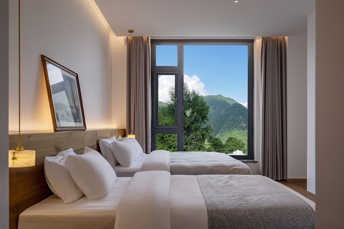 Ein Hotelzimmer mit zwei Betten, einem großen Fenster mit Vorhängen, einem gerahmten Bild an der Wand und einem Nachttisch mit Lampe.