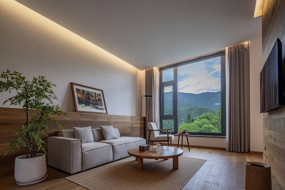 Ein modernes Wohnzimmer mit Sofa, Ohrensessel, Couchtisch und großem Fenster mit Blick auf die Berge.