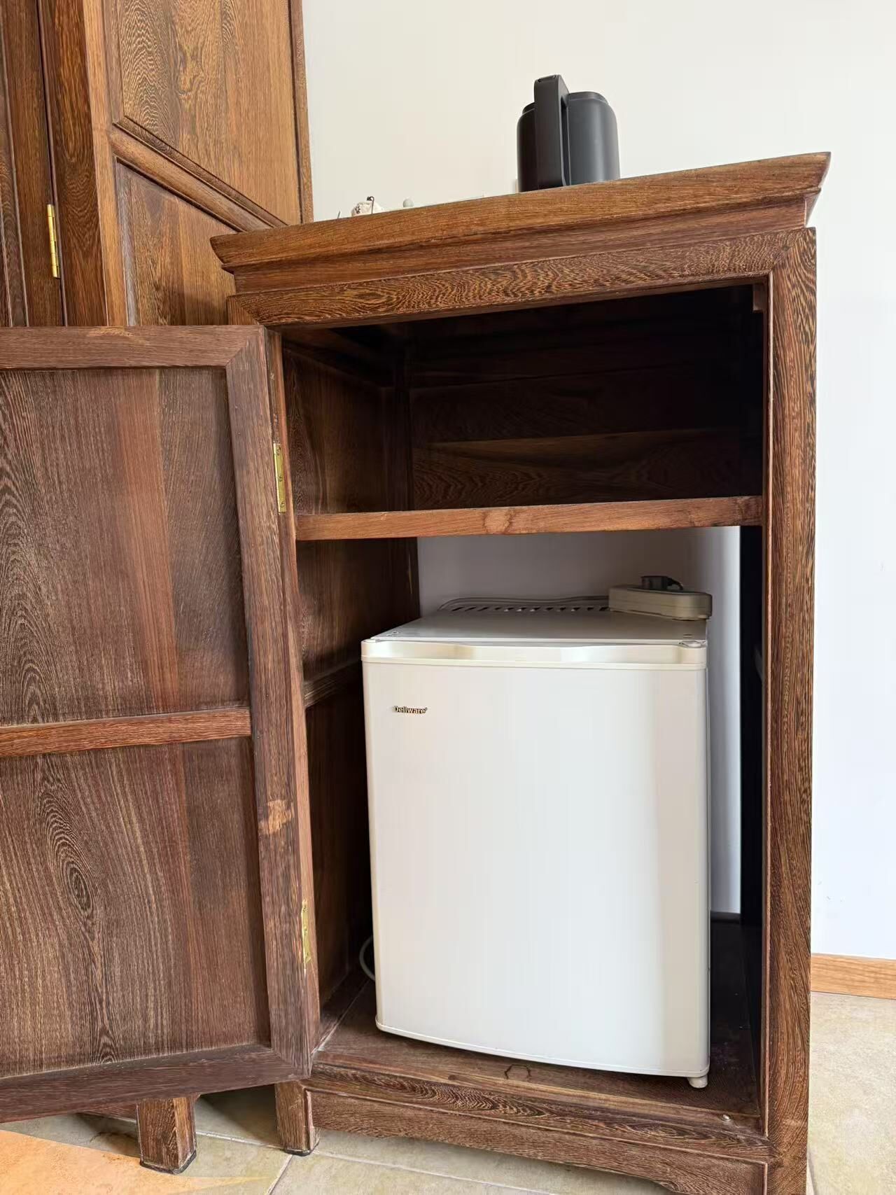 A compact white refrigerator in a wooden cabinet with a shelf above it.