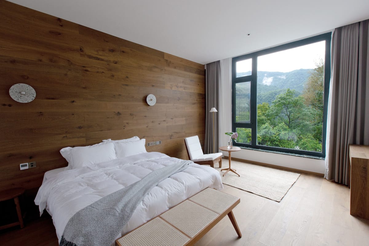 Ein modernes Schlafzimmer mit einem großen Bett, einem Holzkopfteil, Blick auf eine gebirgige Landschaft, einem kleinen Tisch mit Lampe und einem Stuhl.