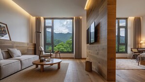 Una sala de estar moderna con sofá, mesa de centro y televisor montado en una pared de madera. Amplios ventanales ofrecen vista a la vegetación y montañas.