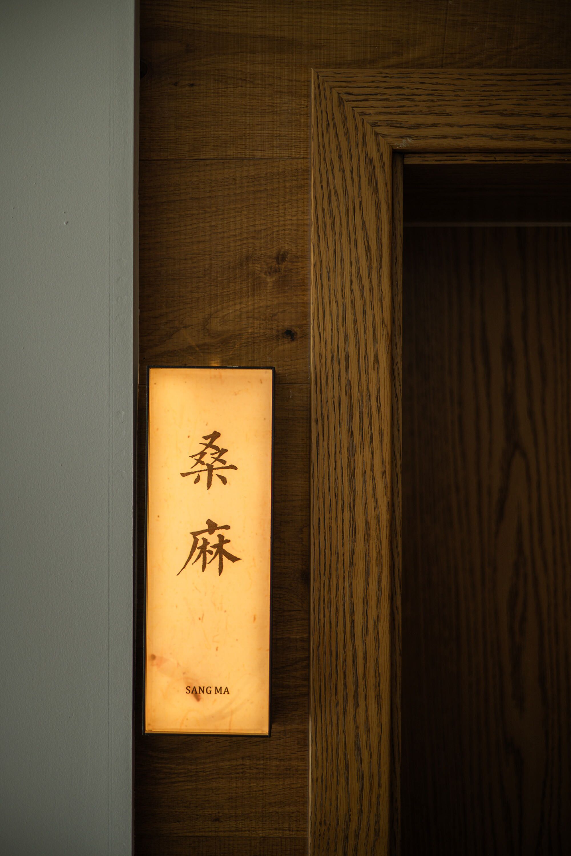 Una puerta de madera con un letrero que muestra caracteres chinos y el nombre 'SANG MA'.