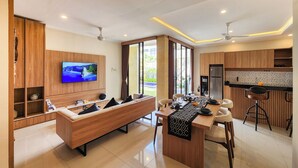 A modern living room with a wooden sofa, a TV mounted on the wall, and a dining area with a kitchen in the background. - Enjoyable Ayur Villa in beautiful Bali (Bali)