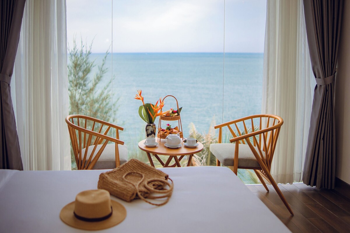 Una habitación con una cama, dos sillones de mimbre, una mesita redonda con una cesta, tetera y tazas, y vista al mar.