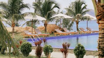 Un espace piscine agrémenté de chaises longues et de parasols, entouré de palmiers et offrant une vue dégagée sur l’eau.