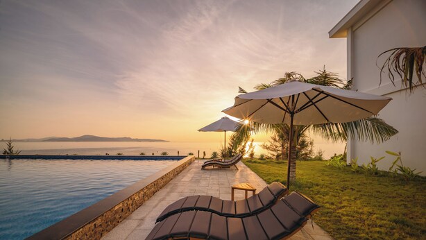 Zona junto a la piscina con sillones de descanso y sombrillas, un muro de piedra y vista al mar.
