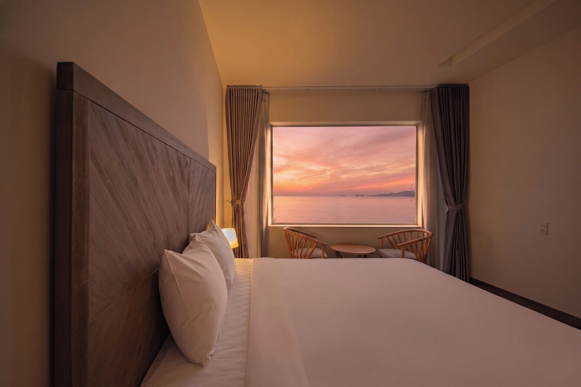 Habitación de hotel con una cama grande, dos sillas, una mesita y vistas al atardecer sobre el agua.
