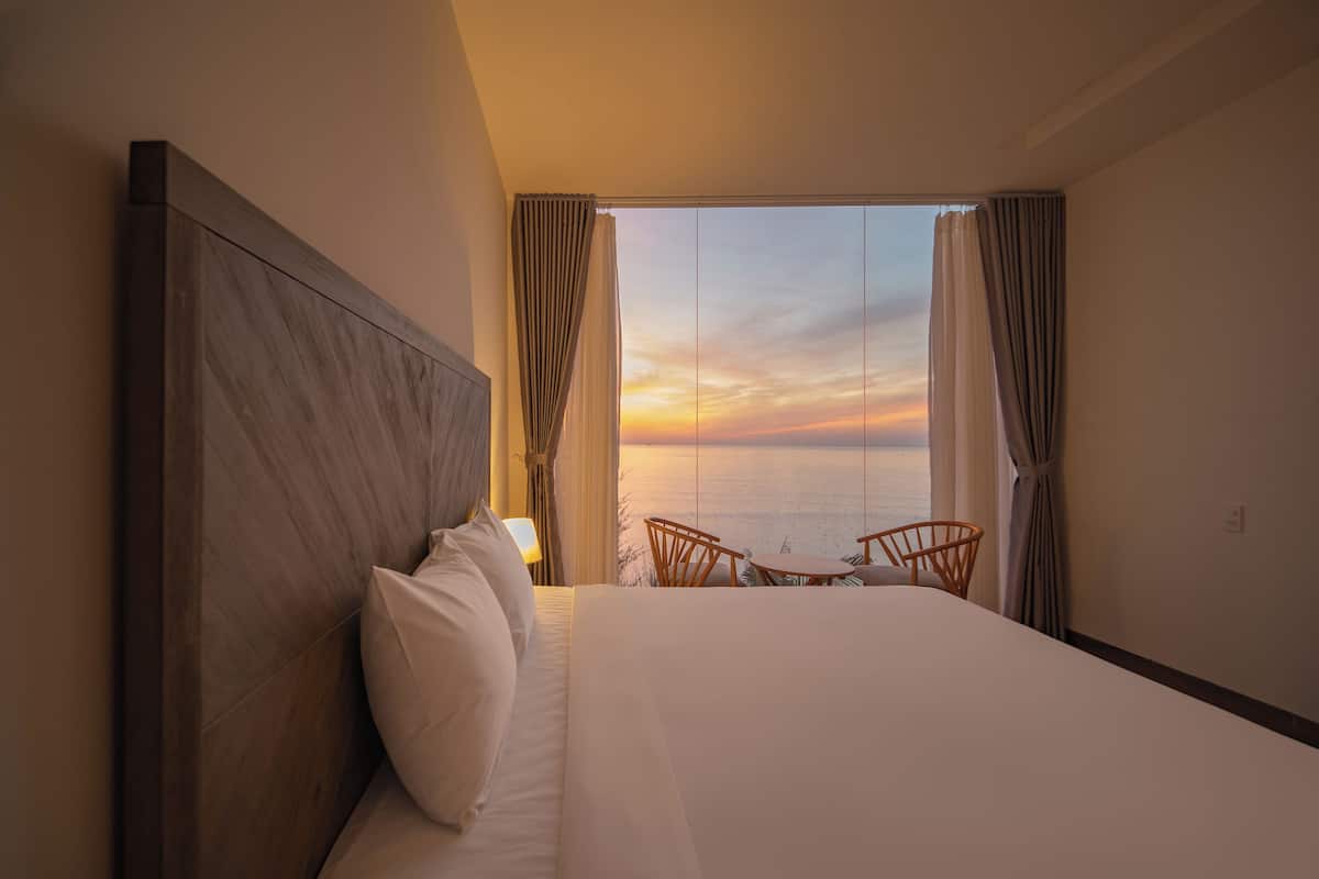 A hotel room with a large bed, a view of the sea, and a small table and chair by the window.