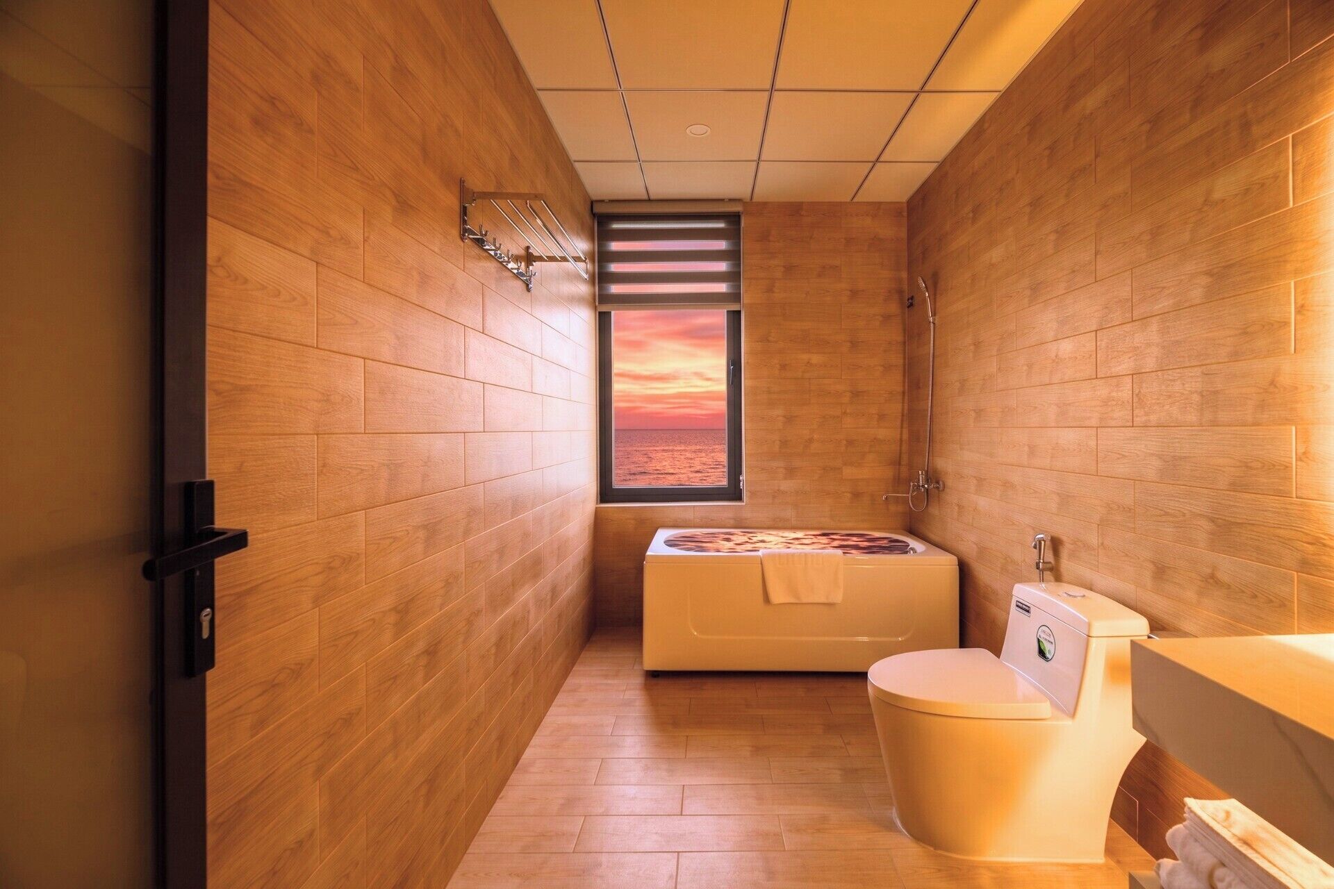 A bathroom with wooden walls, a bathtub, a toilet, and a window with a sunset view.