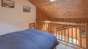 A bedroom with a wooden ceiling, a skylight, a bed with a blue cover, and a wooden railing overlooking a balcony. - Apartment in heart of Morillon Village (Morillon)
