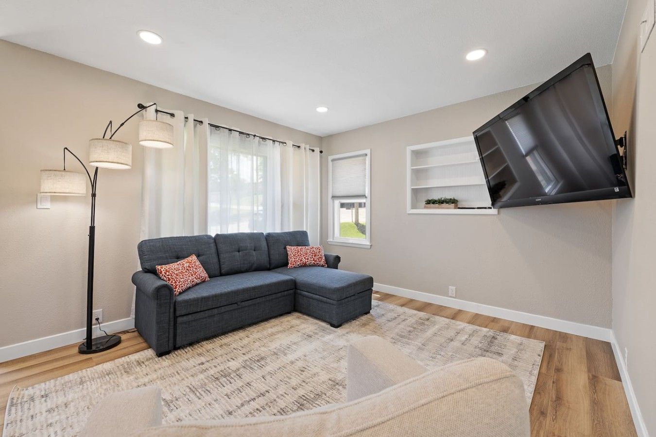 A modern living room with a sectional sofa, a floor lamp, a flat-screen TV mounted on the wall, and a large window with curtains.
