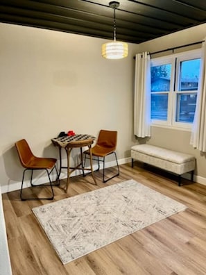A room with a wooden floor, a round table with chairs, a bench, a hanging light fixture, and a window with curtains. - Sleeps 10, Available Garage! (Sturgis)