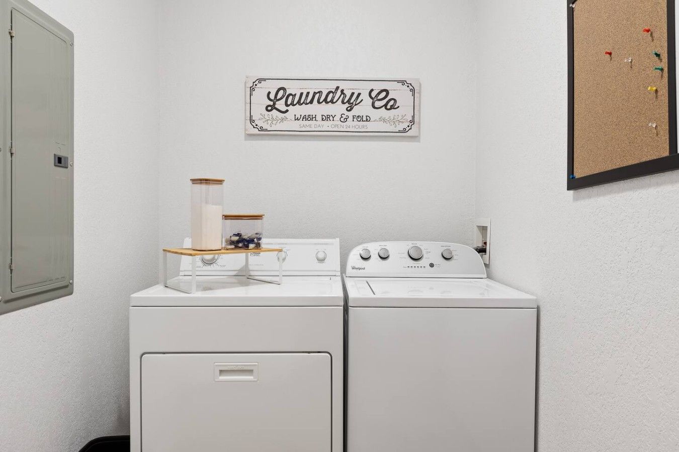 Ein Waschraum mit zwei weißen Waschmaschinen und Trocknern, einem Holzregal mit Gläsern sowie einem Schild mit der Aufschrift 'Laundry Co Wash, Dry & Fold Same Day - Open 24 Hours'.