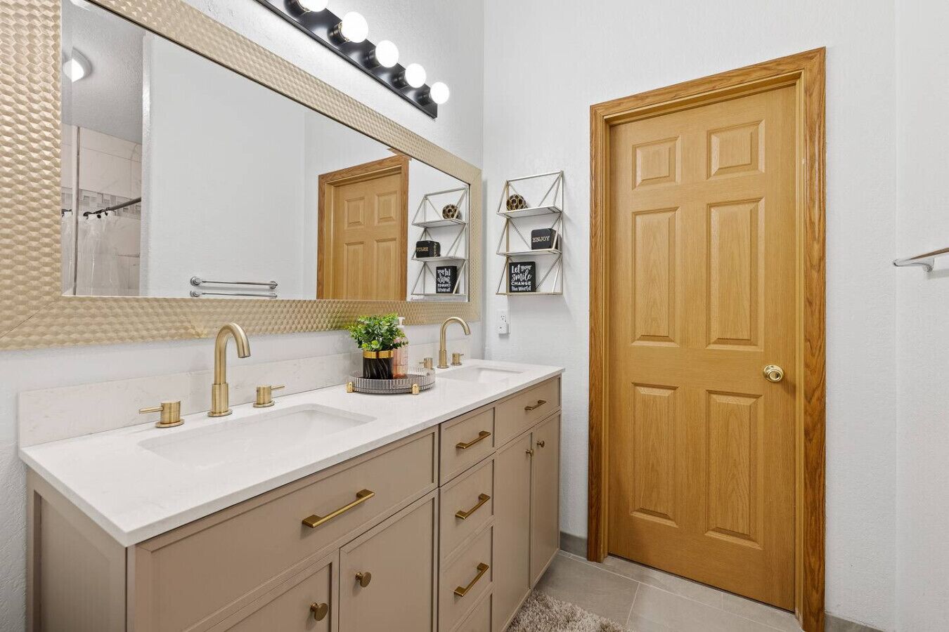A bathroom with a double sink, a large mirror with lights, a wooden door, and a small shelf with decorative items.