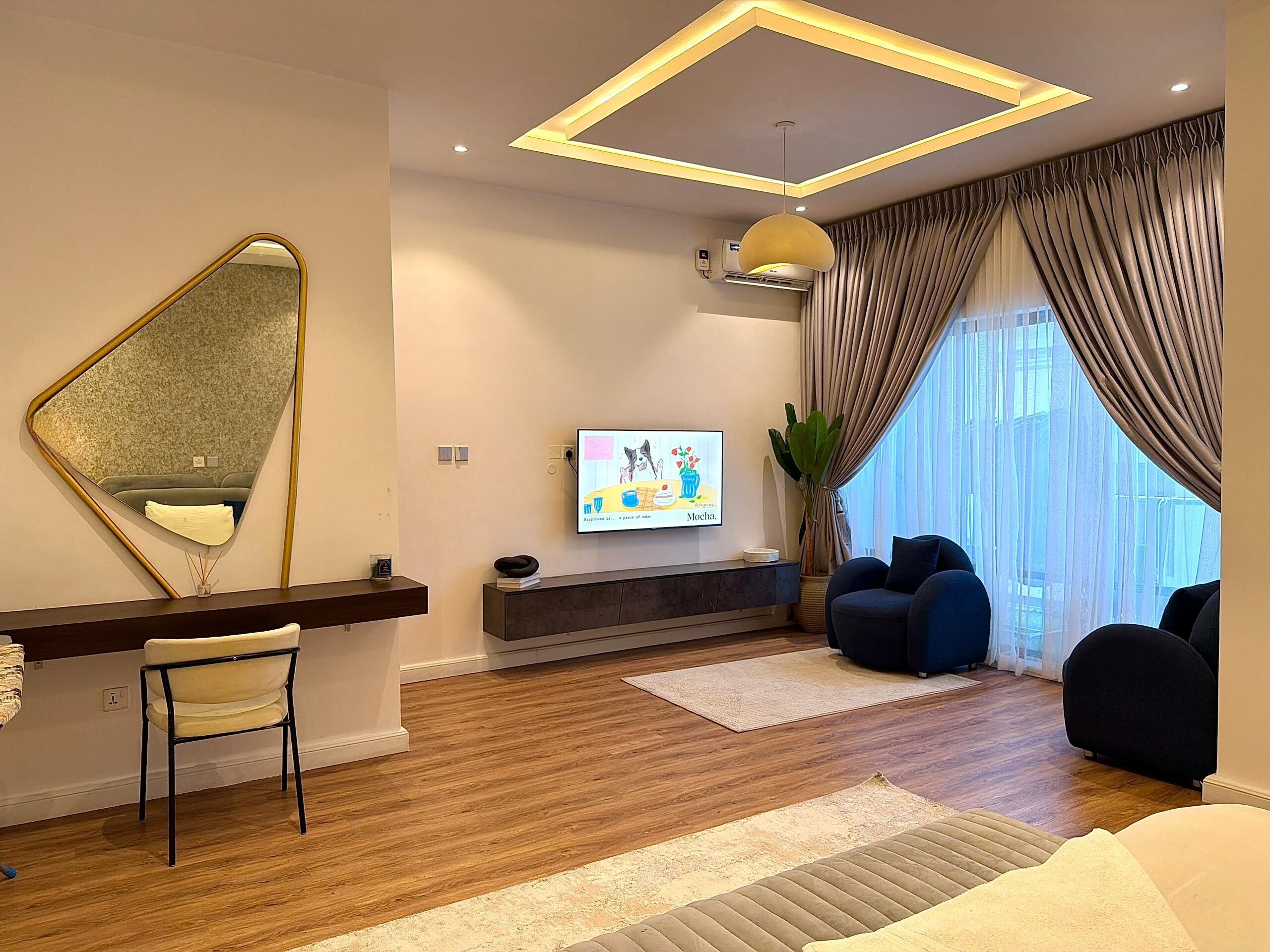 A modern living room with a wooden floor, a large mirror, a flat-screen TV, a sofa, and a dining area with a chair and table.