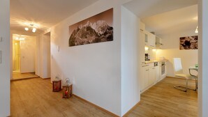 A modern kitchen with white cabinets, a dining area with a white chair, and a hallway with wooden flooring and a mountain painting. - Jagerhaus Apartment Near ski Area (Hopfgarten im Brixental)