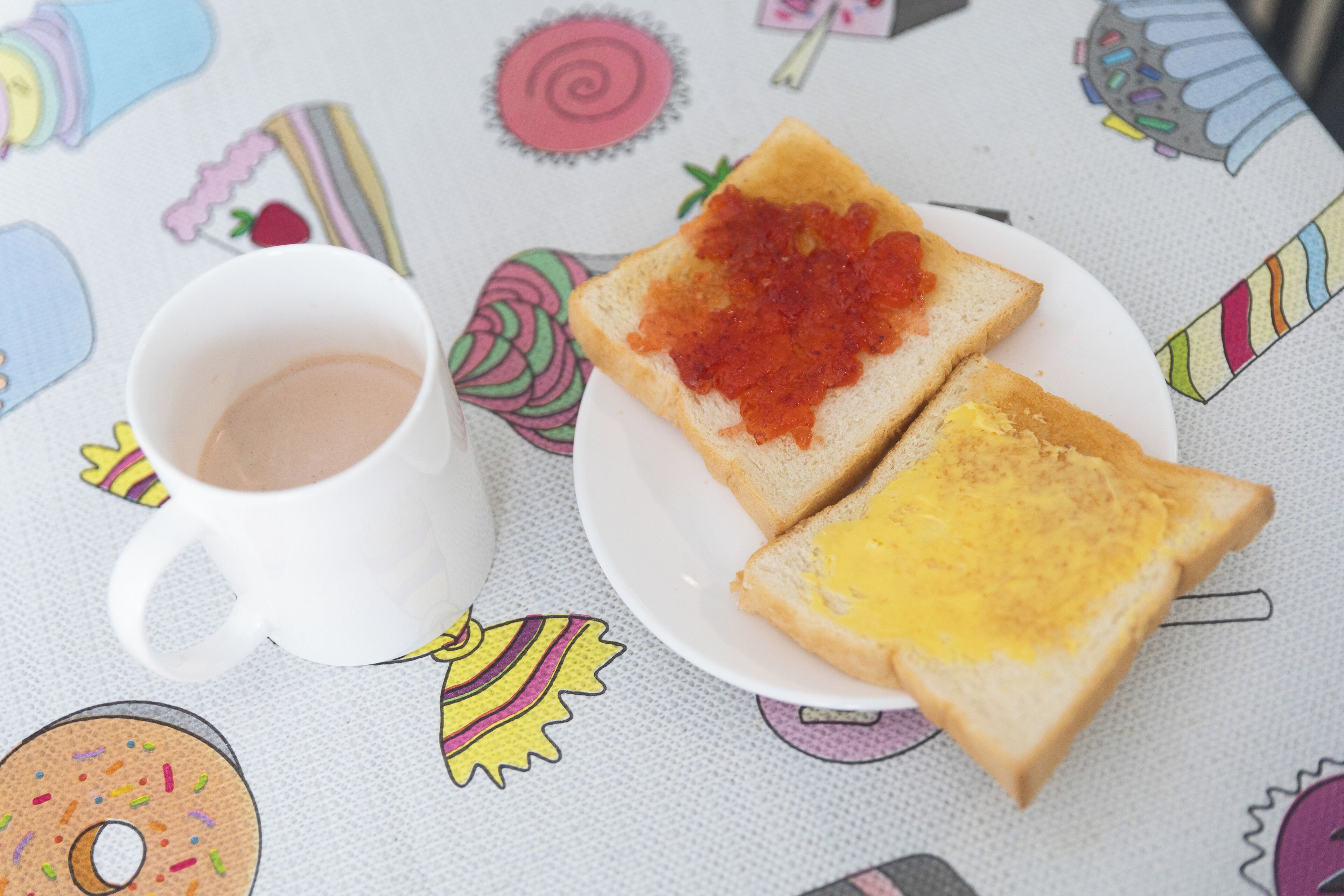 A white mug with a beverage on a table with a colorful tablecloth featuring various food illustrations.