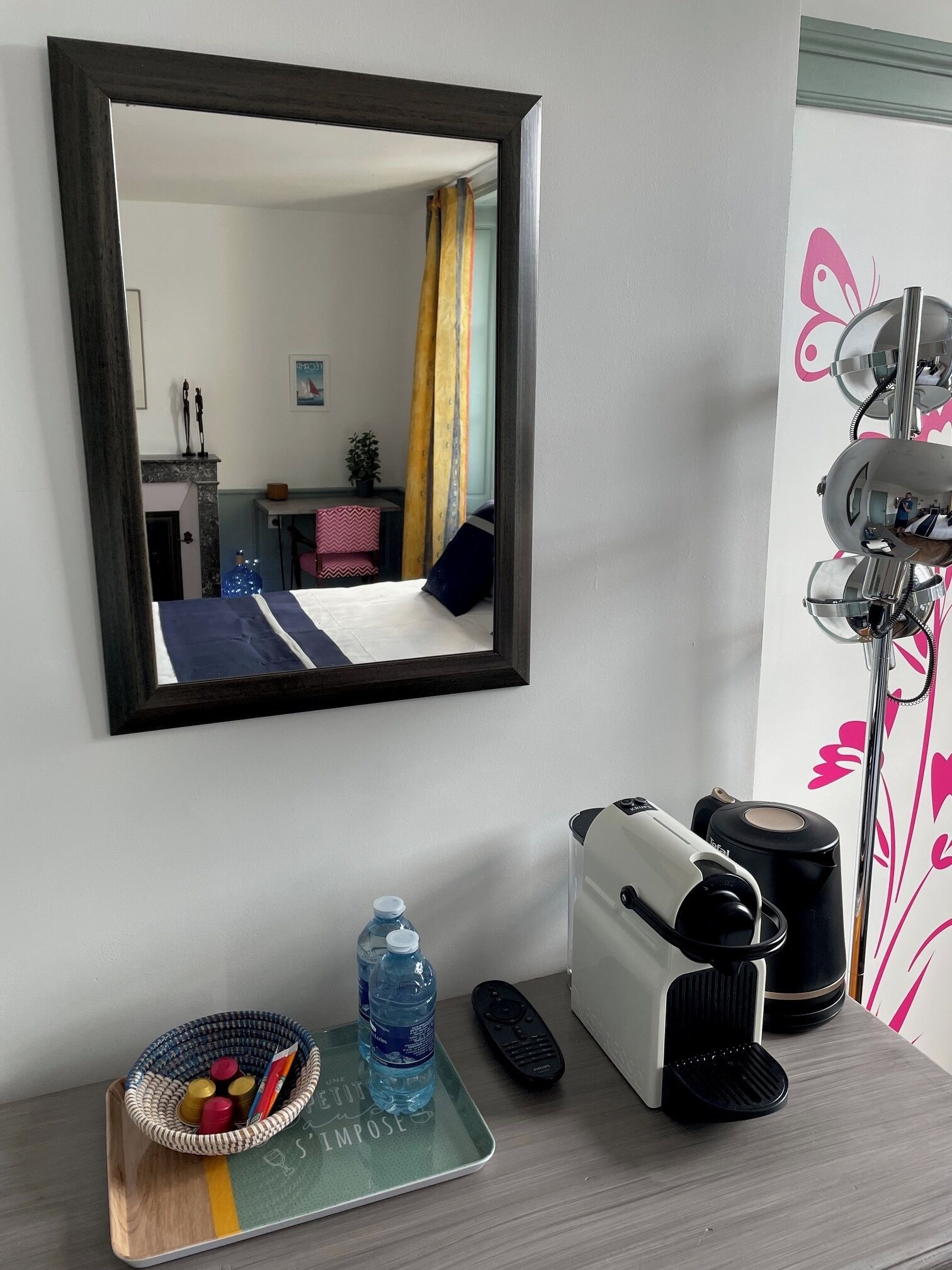 A hotel room with a bed, a desk with a lamp, a mirror, a water bottle, a remote control, and a coffee maker.
