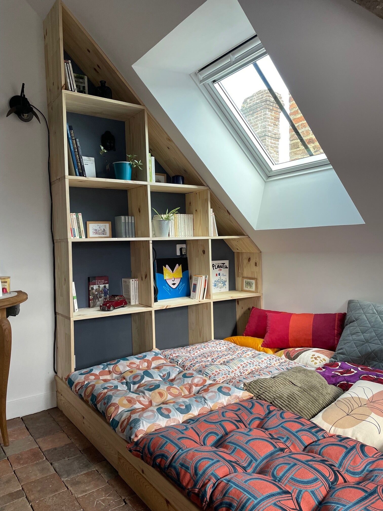 A loft bedroom with a sloped ceiling, a skylight, a built-in wooden bookshelf, a bed with patterned bedding, and a small table with a cup.
