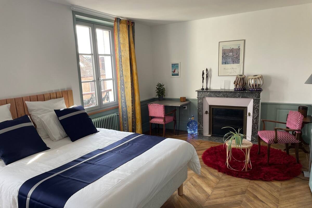 Une chambre avec un grand lit, une cheminée, un bureau et une vue sur l’extérieur.