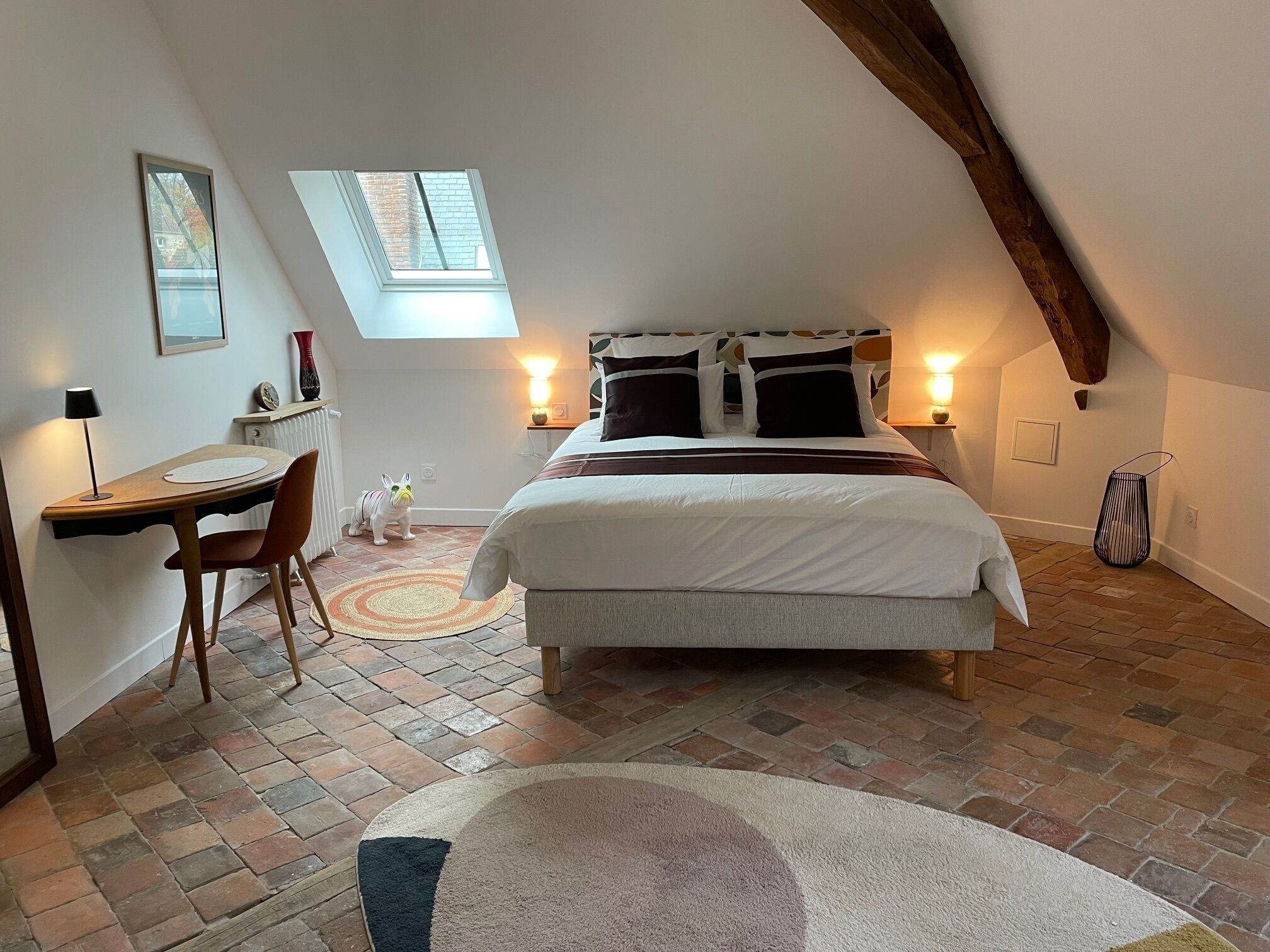 A cozy attic bedroom with a large bed, a desk, and a chair. There is a skylight window and a small table with a lamp.