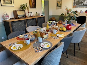 Mesa de comedor preparada con alimentos para el desayuno, rodeada de sillas, en una habitación con chimenea y decoración en la pared.