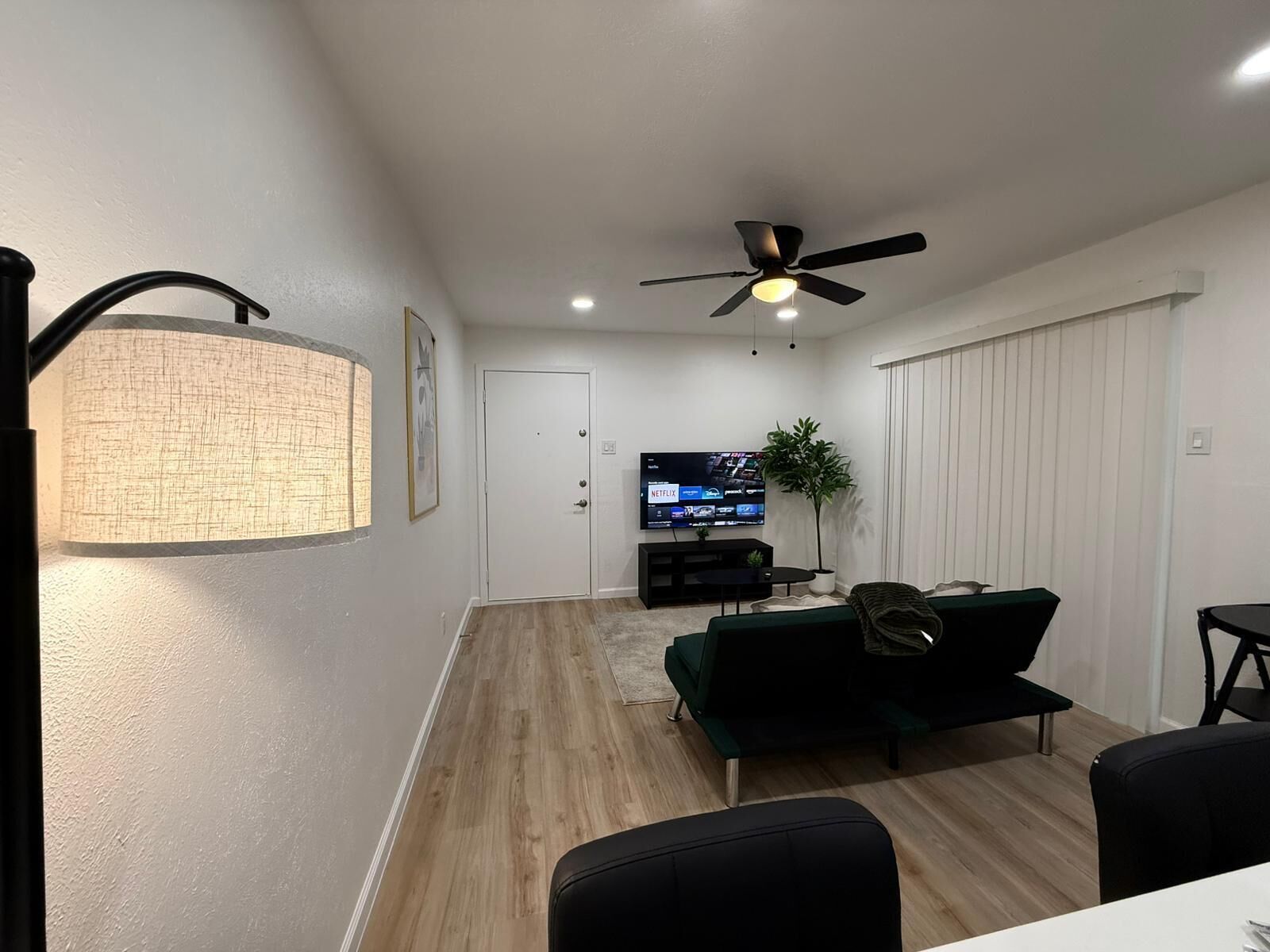 A modern living room with a flat-screen TV, a ceiling fan, a dining area with a table and chairs, and a large window with blinds.