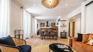 A modern living room with a sofa, a coffee table, and a dining area with chairs and a kitchen in the background.