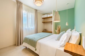 Quarto com cama, mesa de cabeceira, luminária pendente, janela com cortinas e prateleira com toalhas.