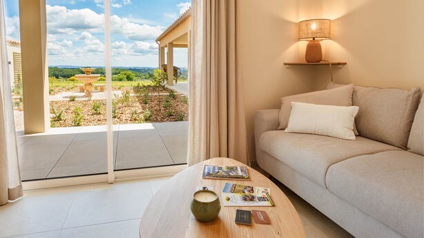 A room with a sofa, a table with a candle and magazines, a lamp, and a view of a garden and building through large windows. - Domus Olivea (Estézargues)