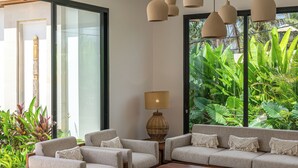 A modern living room with a sectional sofa, ottomans, a coffee table, pendant lights, and a view of lush greenery through large windows. - Villa Anarya Ubud (Ubud)
