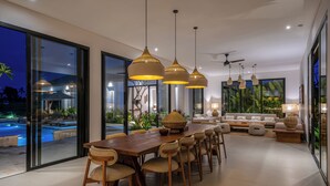 A modern dining area with a long wooden table and chairs, pendant lights, and a view of an outdoor swimming pool. - Villa Anarya Ubud (Ubud)